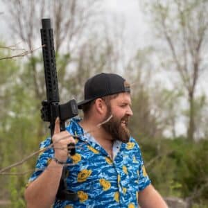 A man in a floral shirt holds a black rifle, smiling outdoors in a natural setting, showcasing American craftsmanship.