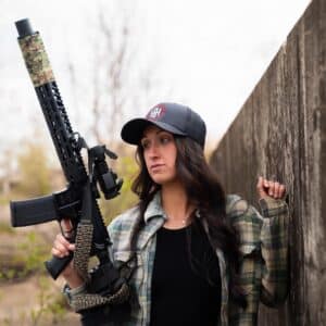 A woman in a flannel shirt holds a reliable American-made rifle featuring a camouflage suppressor wrap, optic sight, and custom paracord sling.