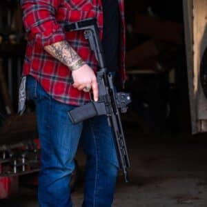 A person in a red plaid shirt holds a reliable, American-made black rifle equipped with a tactical optic and a polymer magazine in a workshop setting.