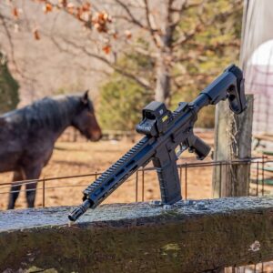 A black American-made rifle with an EOTech optic rests on a wooden fence, with a horse in the background, showcasing reliable, high-quality design.