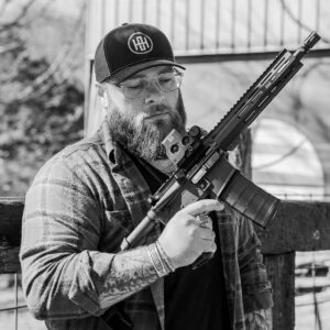 A bearded man wearing a One Horse cap holds a reliable American-made rifle equipped with an EOTech optic, showcasing quality craftsmanship outdoors.
