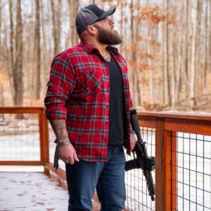 A bearded man in a red flannel shirt stands on a wooden deck holding a reliable, American-made black rifle in a wooded, winter outdoor setting.