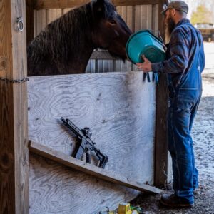A black American-made rifle with an optic rests on a wooden stall wall while a man in denim overalls feeds a horse from a teal bucket nearby.