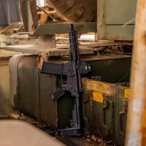 A reliable black American-made rifle with an EOTech optic, resting against a rugged, weathered green military vehicle in a rustic outdoor setting.