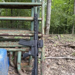 A reliable black American-made rifle with a modular handguard and optic, leaning against a weathered green metal farm gate in a wooded setting.