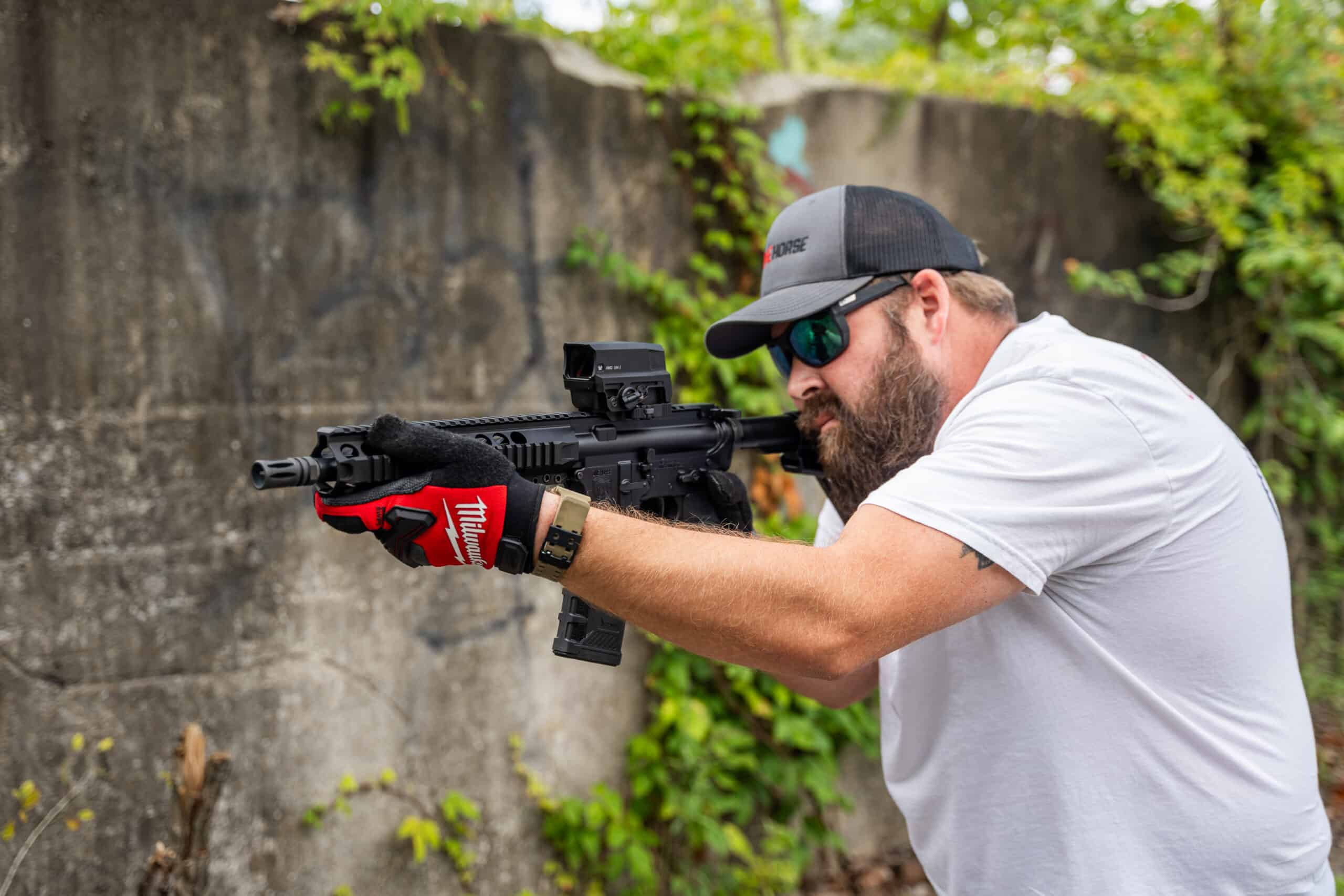 A bearded man in a One Horse cap aims a black American-made rifle with a red dot sight, wearing protective gloves against a weathered concrete wall.