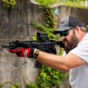 A bearded man in a One Horse cap aims a black American-made rifle with a red dot optic, wearing red and black work gloves for a reliable grip.
