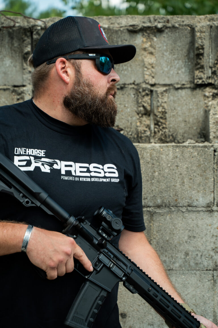 Man in a One Horse Express t-shirt holding a black One Horse Express rifle by a stone wall