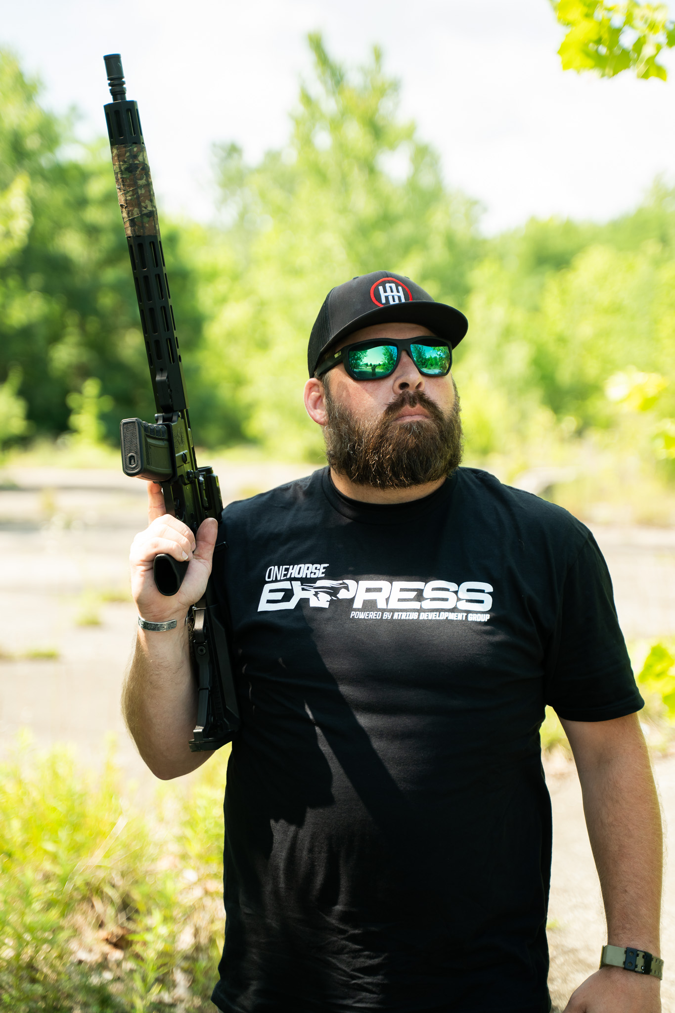 Man holding a camouflaged rifle outdoors, wearing sunglasses and a One Horse Express t-shirt, showcasing American craftsmanship.