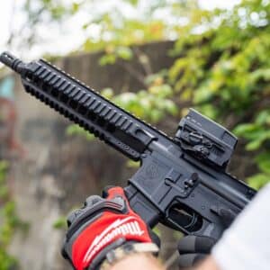 A reliable One Horse Model GH-15 rifle with a black finish, featuring a tactical rail and mounted optic, held by a person wearing red work gloves.