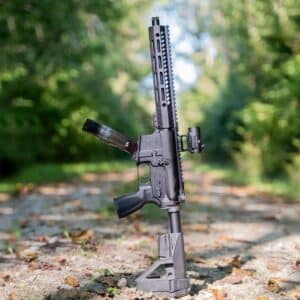 A reliable American-made One Horse rifle with a black finish, M-LOK handguard, Vortex optic, and drum magazine, standing upright on a forest trail.