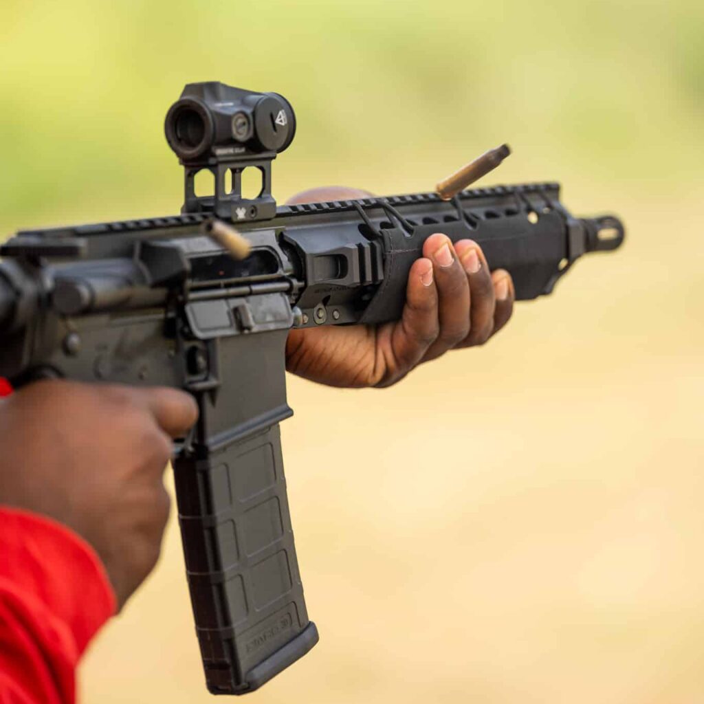 Close-up of a black AR-15 with a red dot sight and polymer magazine as a spent brass case leaves the ejection port
