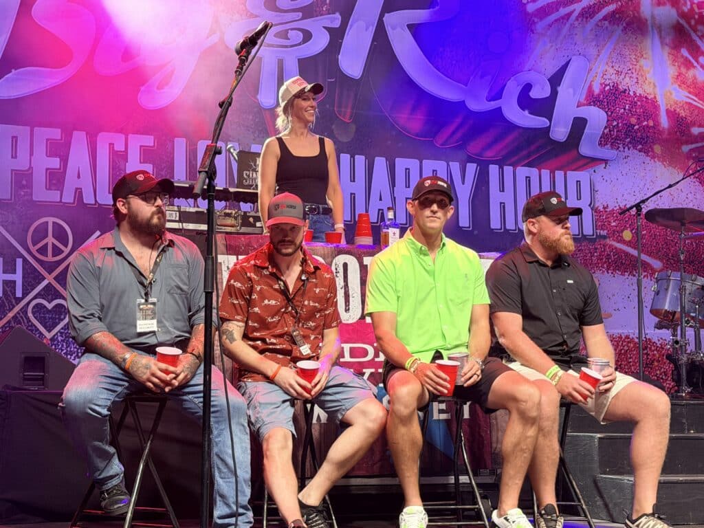 Four men and one woman wearing One Horse branded hats sit on stage at a Big and Rich concert, promoting American-made quality and reliability.
