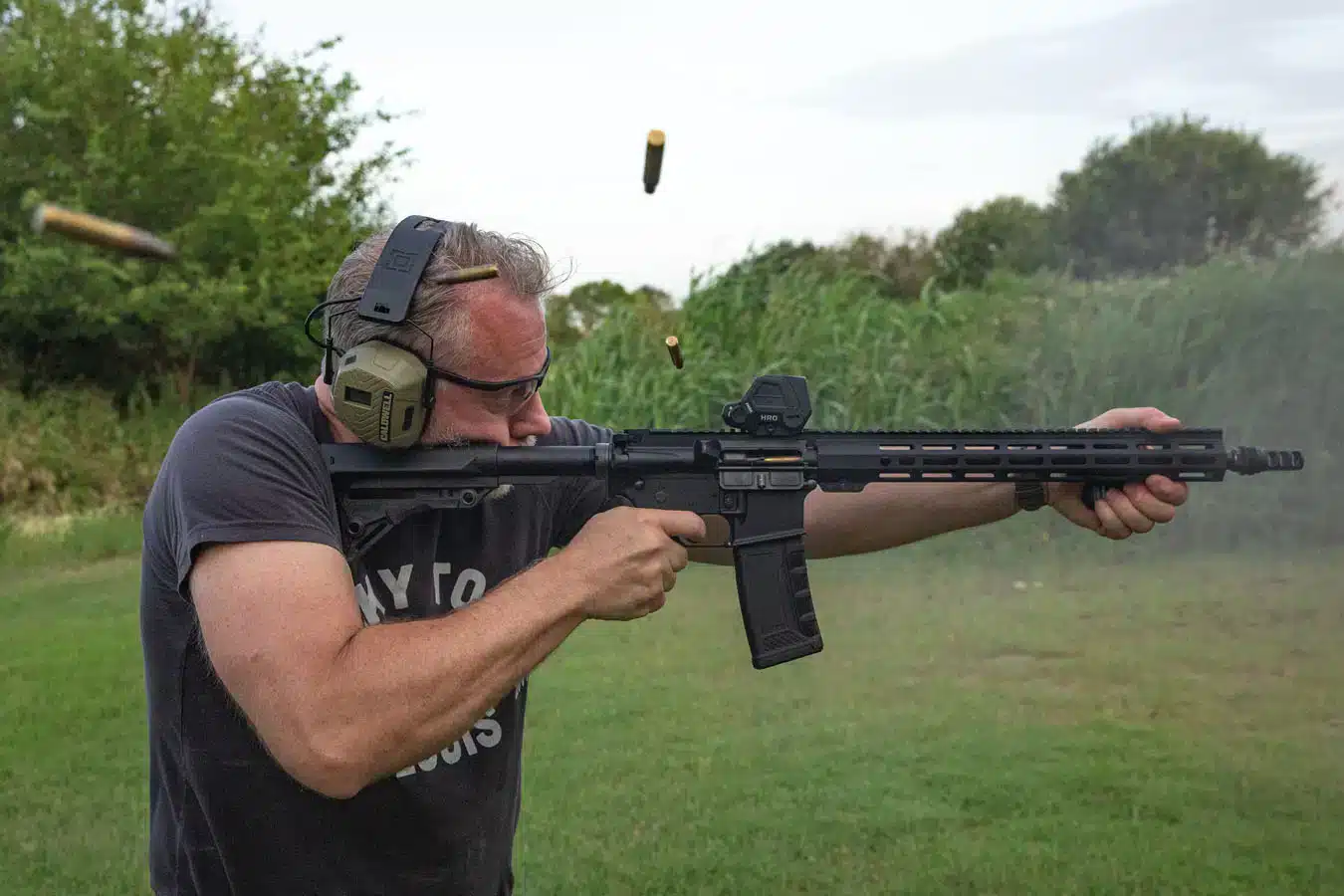 A shooter firing a black American-made AR-15 rifle with an optic at an outdoor range, showing reliable performance with spent casings ejecting.