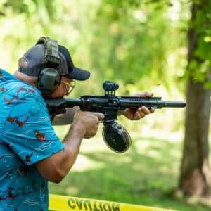 A shooter wearing ear protection fires a black rifle with a drum magazine and red dot sight at an outdoor range, with brass casings ejecting mid-air.