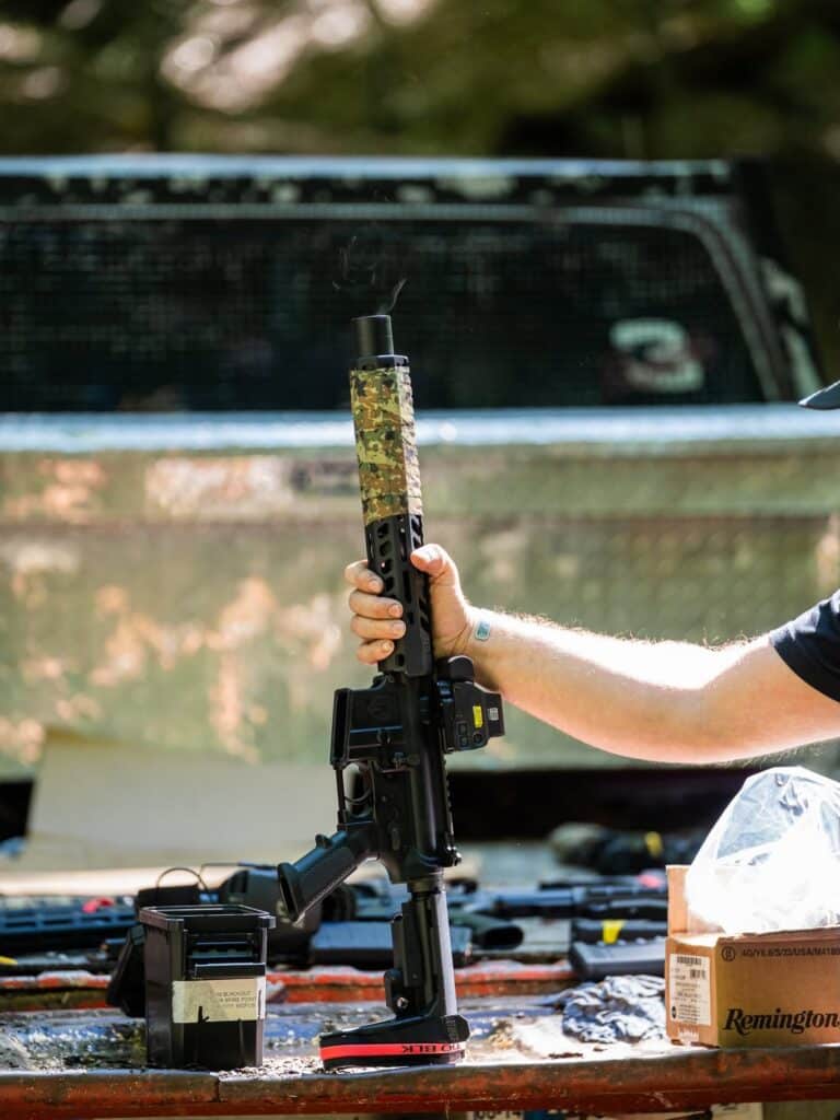 Shooter holding an AR-15 with a camo-wrapped handguard and holographic sight, smoke rising after a string of shots